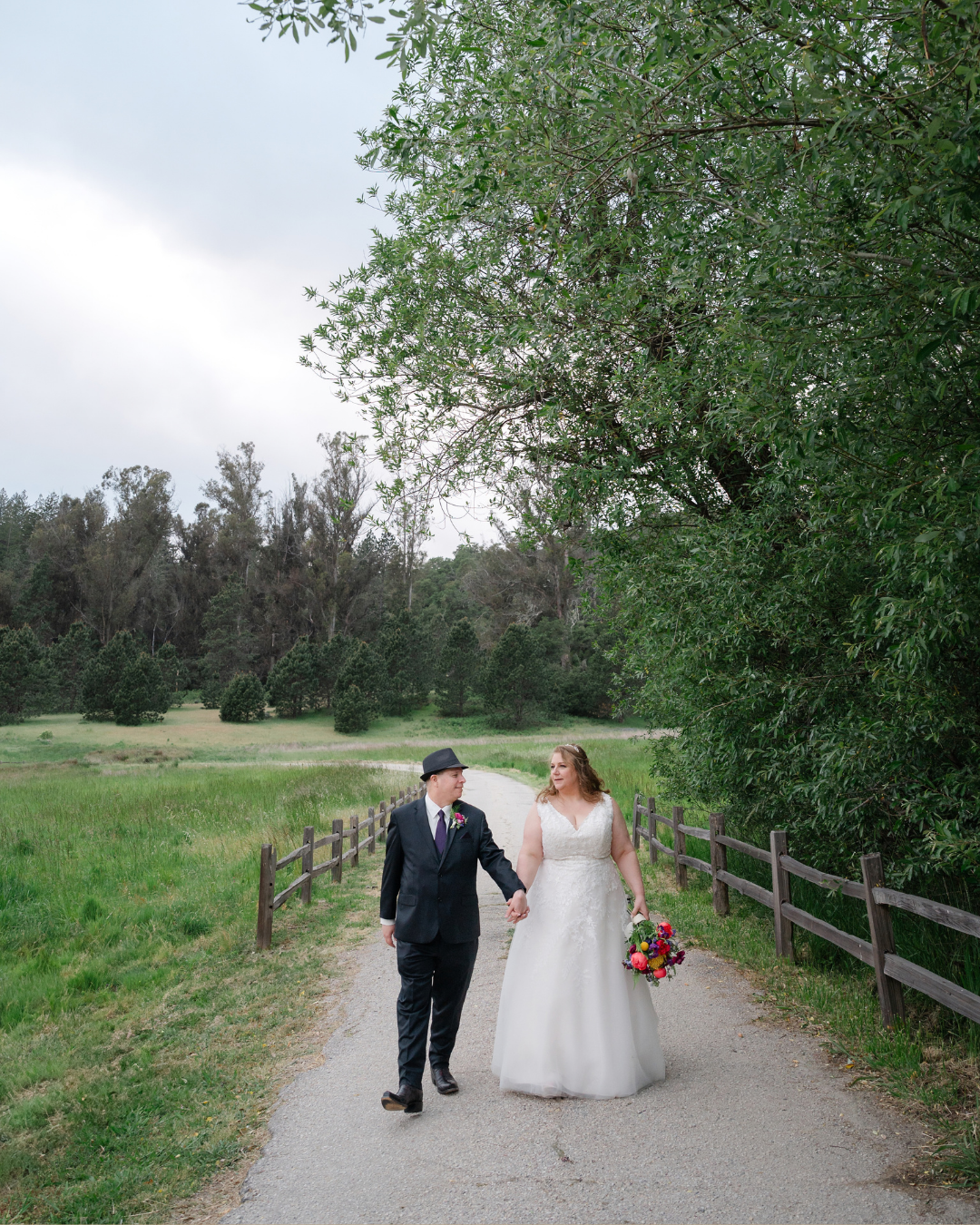 A bride and groom walk hand-in-hand down a scenic path surrounded by lush green trees and a wooden fence.