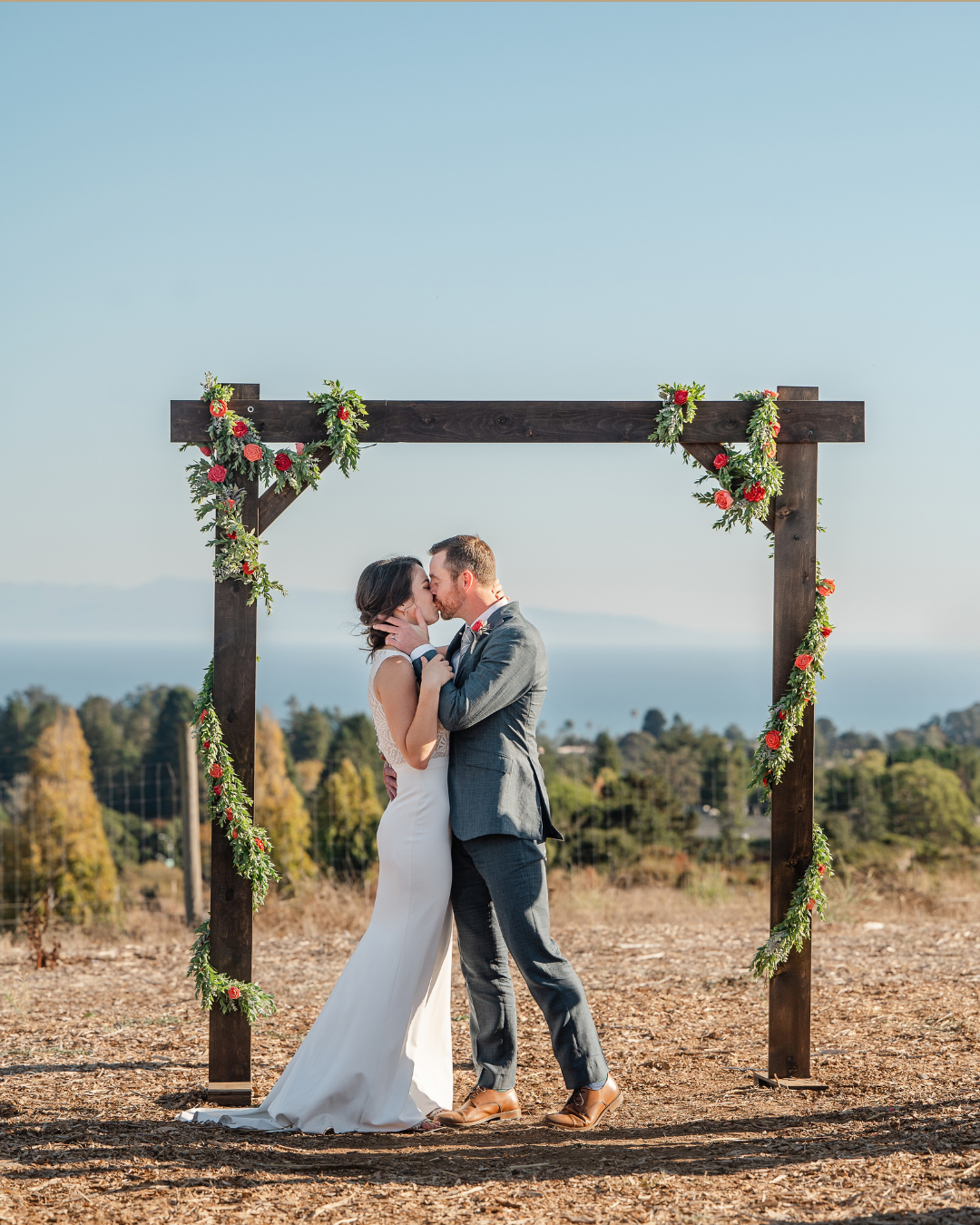 Bride and groom share first kiss under rustic wooden arch adorned with greenery and red flowers with ocean views behind.