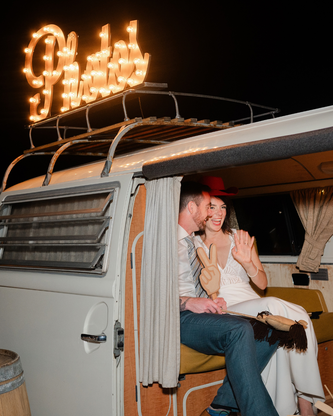 Couple laughing in vintage VW bus with glowing marquee sign on roof at night wedding celebration.