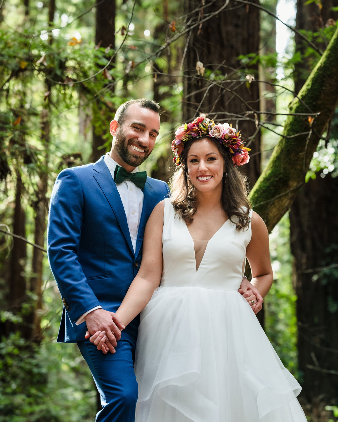 Bride and groom smiling in redwood forest, bride wearing floral crown and white gown, groom in blue suit.