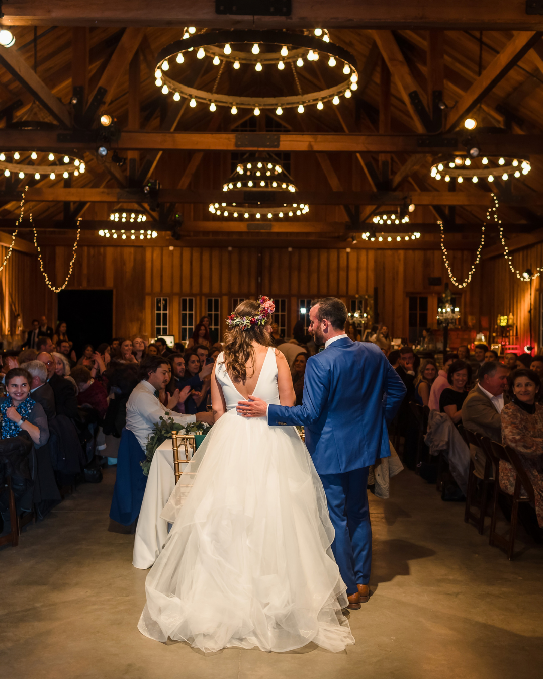 Bride and groom share first dance in rustic barn wedding venue with warm string lights and circular chandeliers.