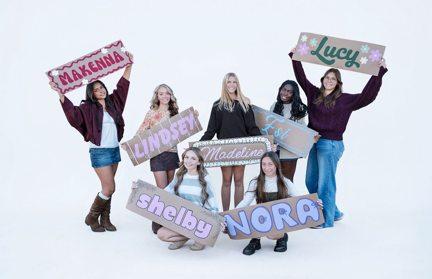 Seven young women pose together holding colorful personalized name signs reading Makenna, Lindsay, Lucy, Madeline, Shelby, and Nora.