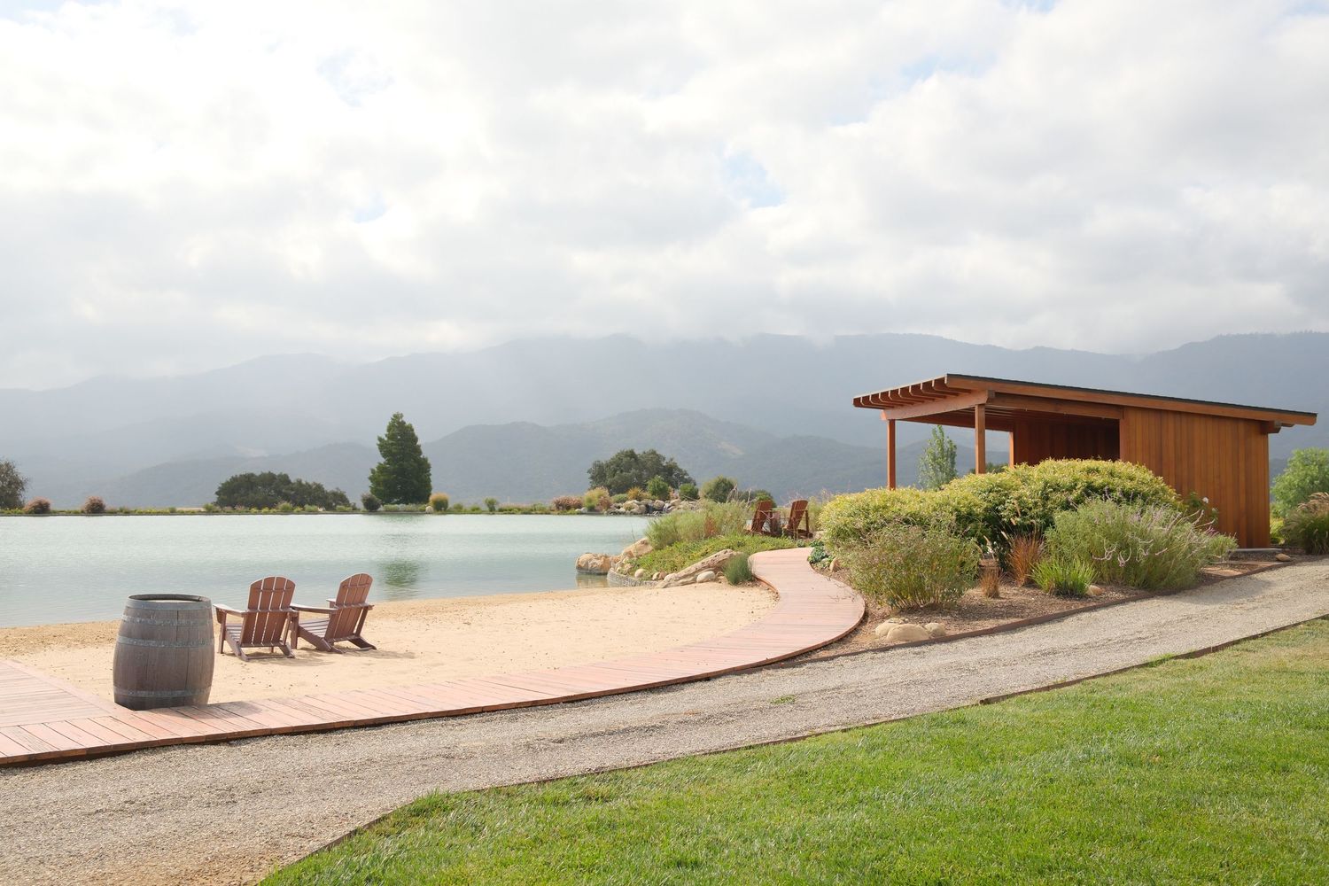 Scenic winery pond with Adirondack chairs, wooden barrel, pergola, and mountain views under cloudy skies.