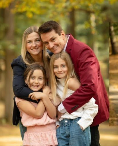 Happy family of four posing outdoors in autumn park, parents hugging two blonde daughters in warm fall colors.