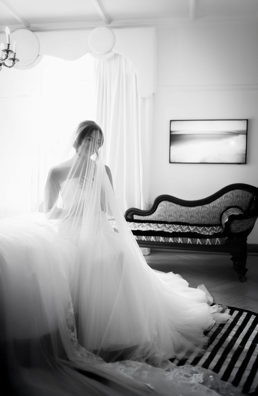 Bride in flowing white gown and veil stands by bright window in elegant room with vintage chaise lounge.
