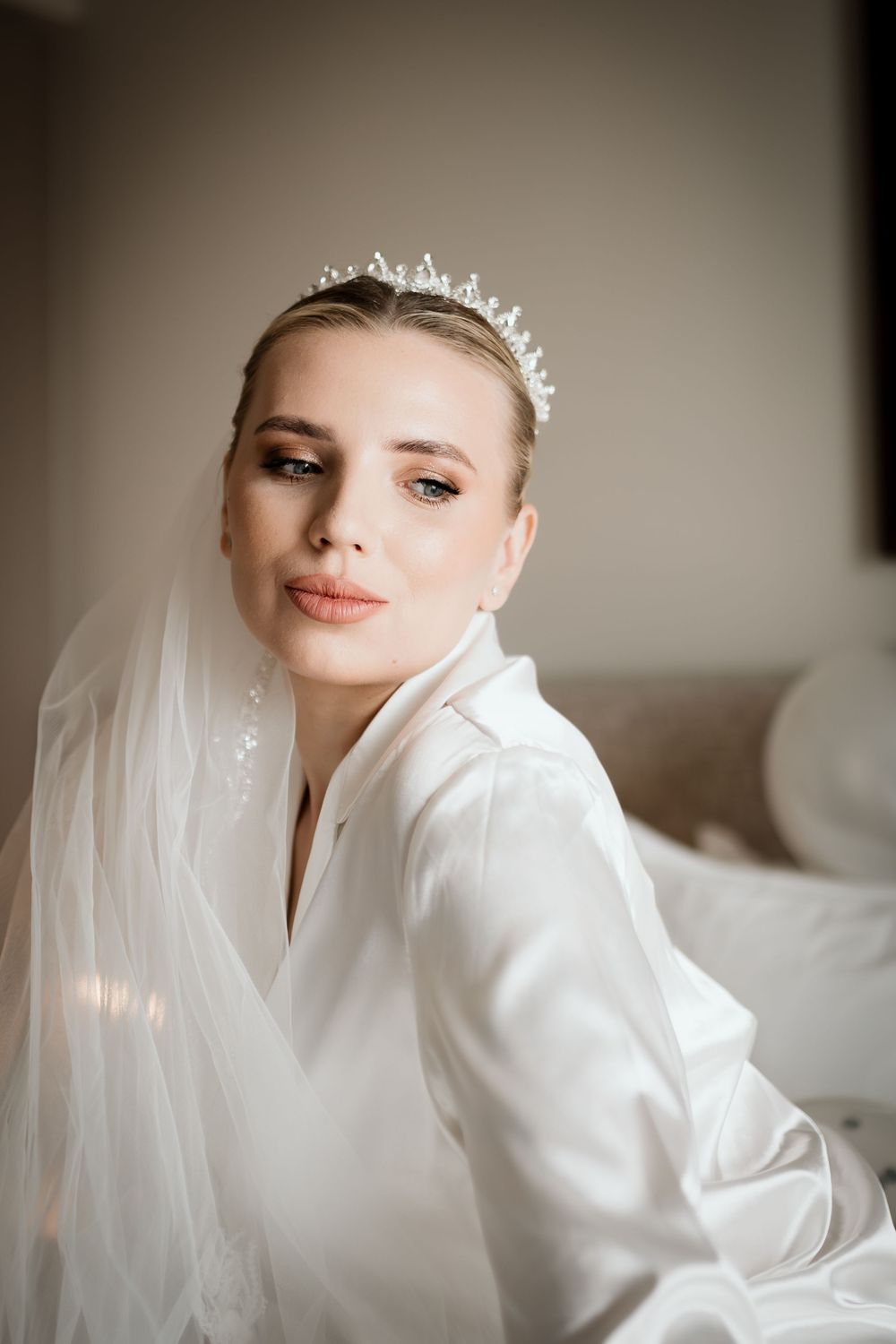 Elegant bride wearing a crystal tiara and white satin robe with a flowing veil, posing gracefully for bridal portrait.