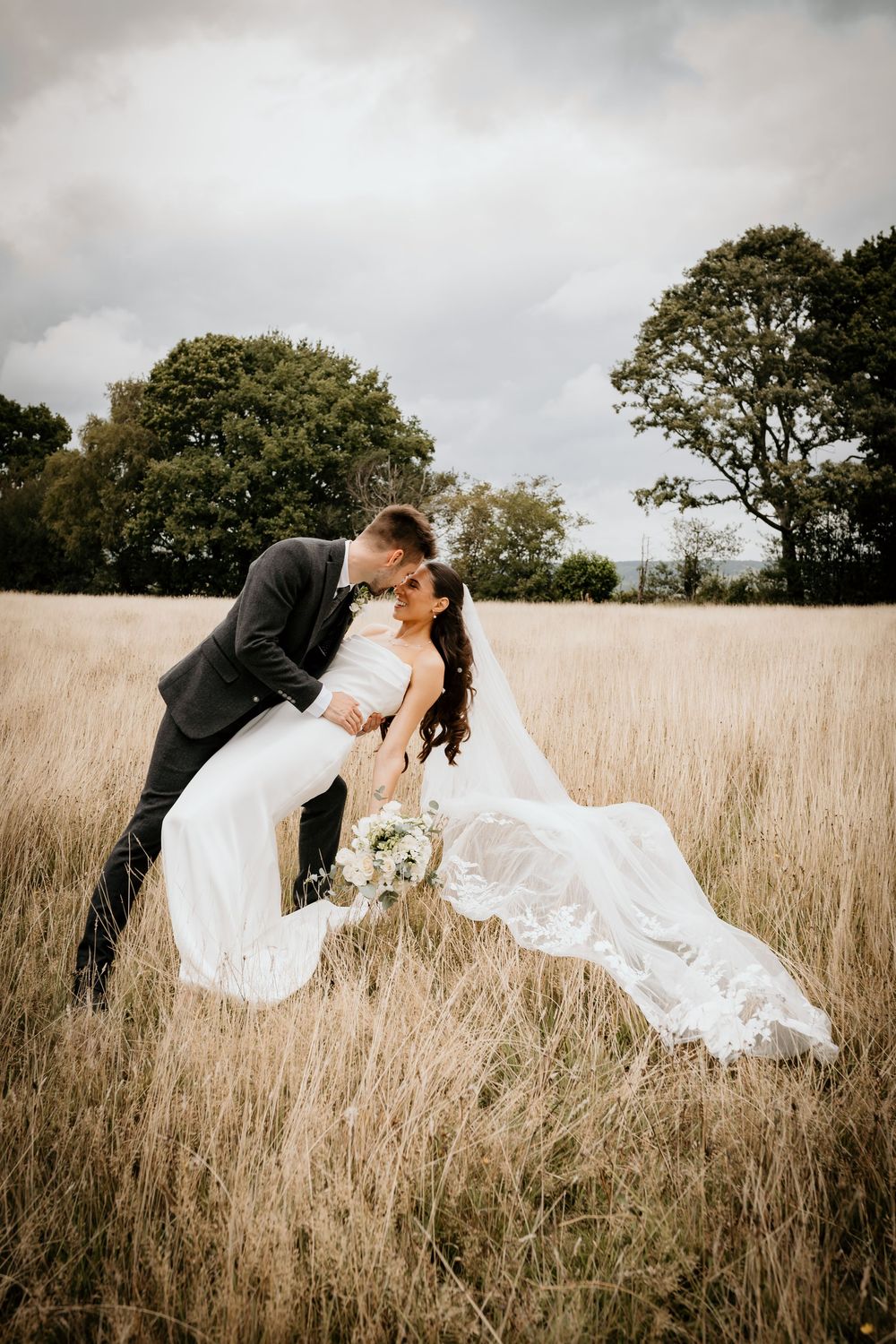 A groom dips his bride for a kiss in a golden wheat field, her white gown and veil flowing, with trees and cloudy sky behind.