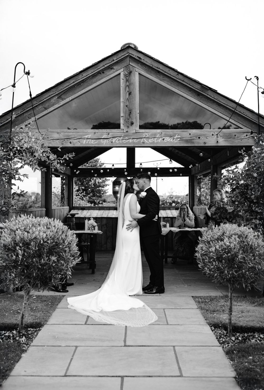Black and white photo of a bride and groom sharing a kiss under a rustic wooden pergola on their wedding day.