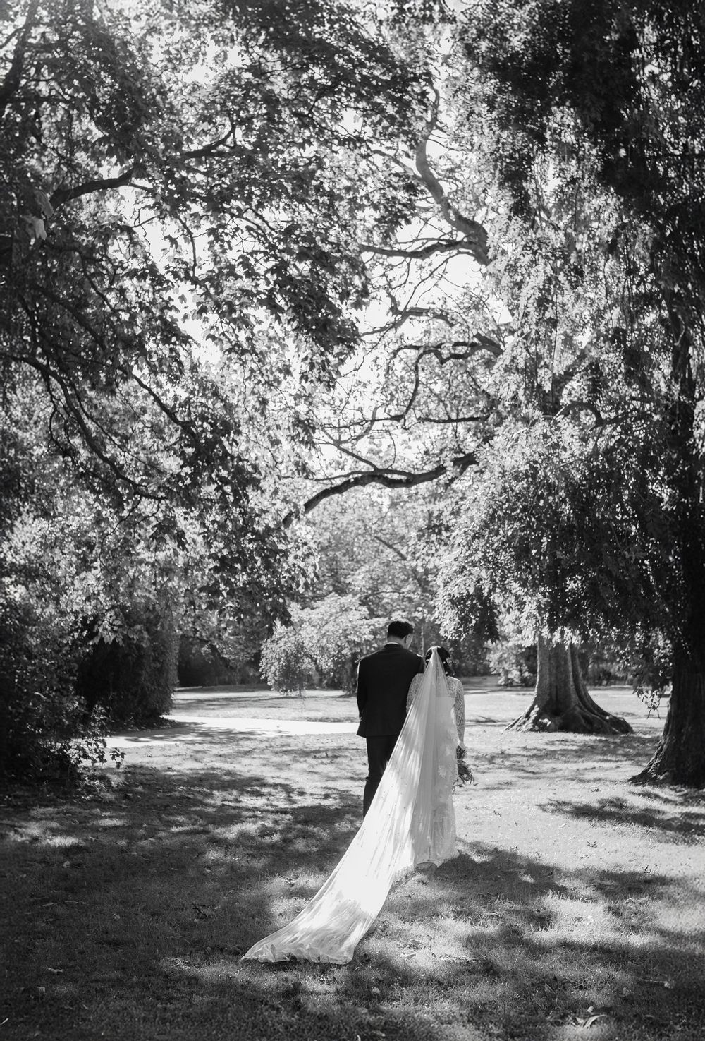 Black and white photo of a bride and groom walking through a tree-lined park path on their wedding day.