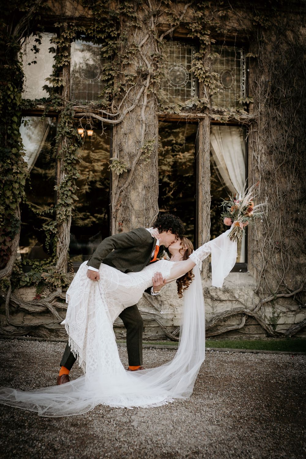 Groom dips bride in a romantic kiss outside a rustic ivy-covered venue, bride in lace gown holding floral bouquet.