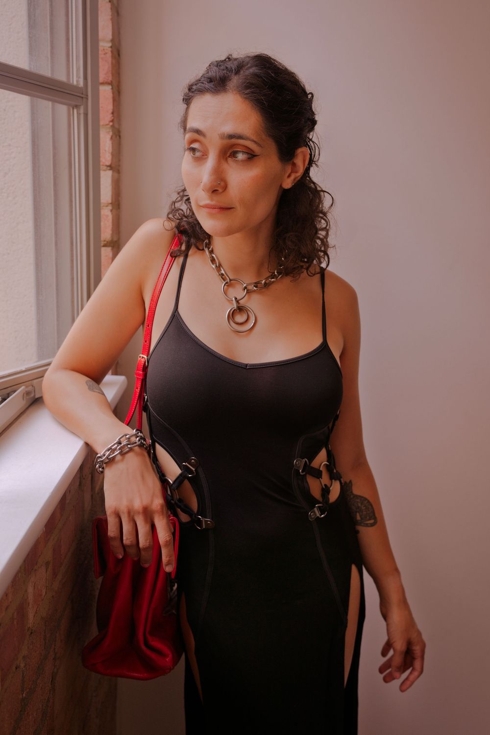 Woman in black cutout dress with chain necklace and red handbag posing near window.