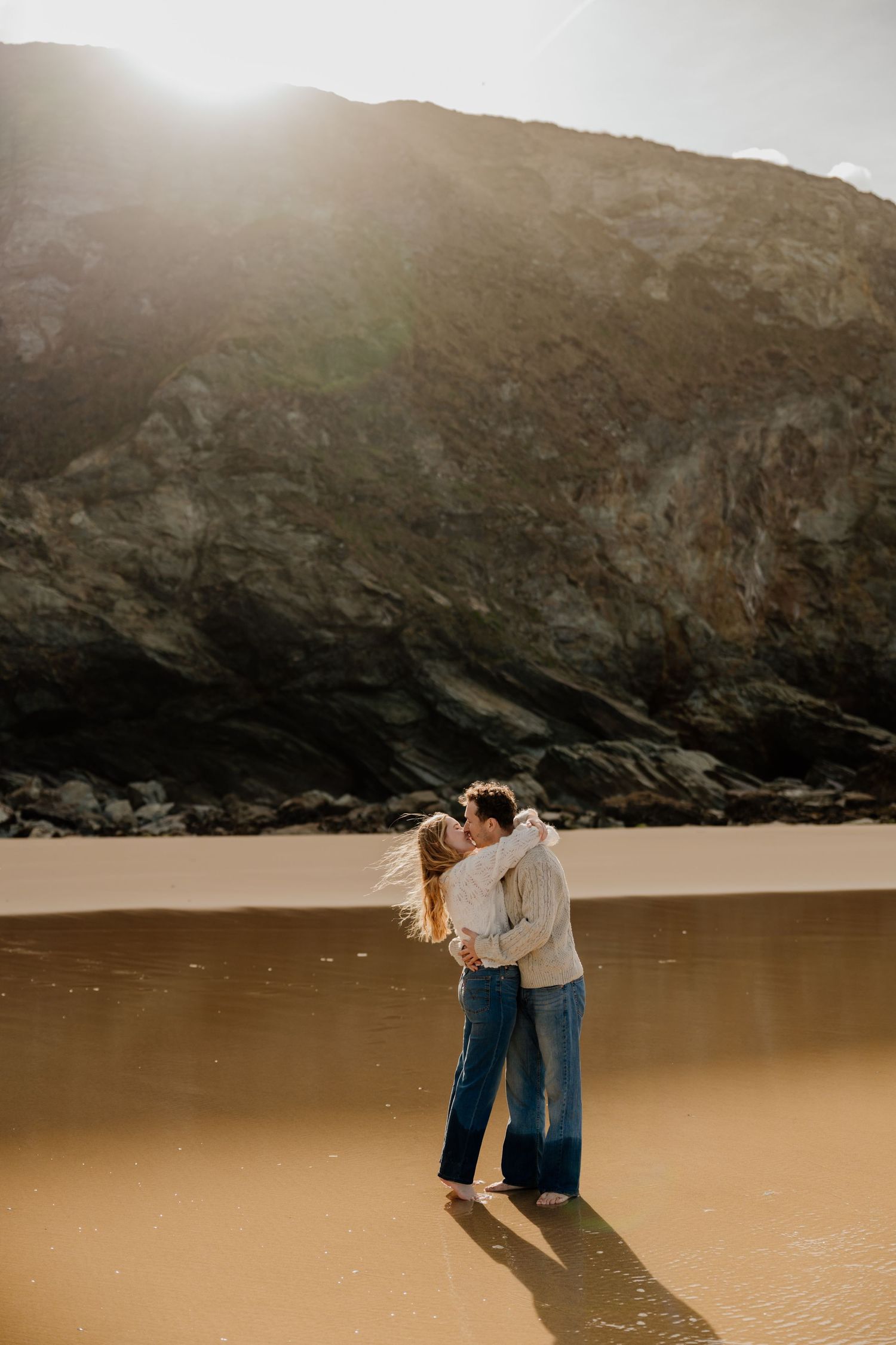 Couple embracing and kissing at watergate bay with dramatic rocky cliffs and golden sunlight behind them.