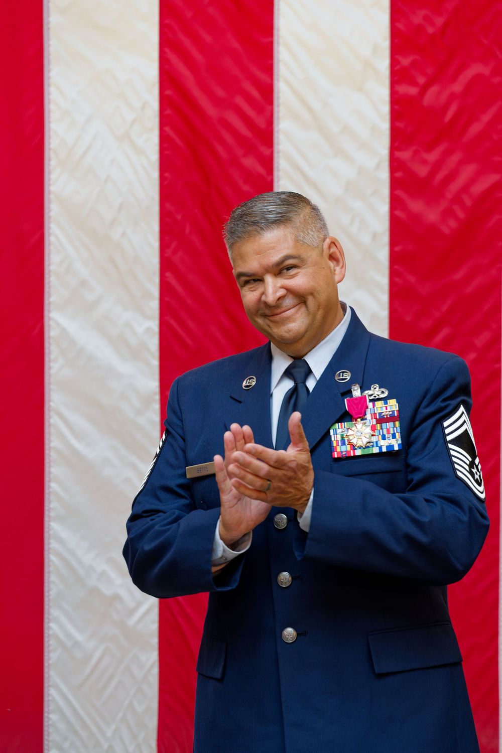 Smiling Air Force Chief Master Sergeant applauds in dress blues before an American flag.
