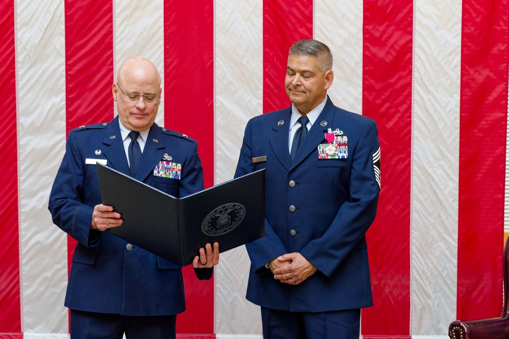 Two Air Force officers stand before American flag, one reading from a certificate during a military ceremony.