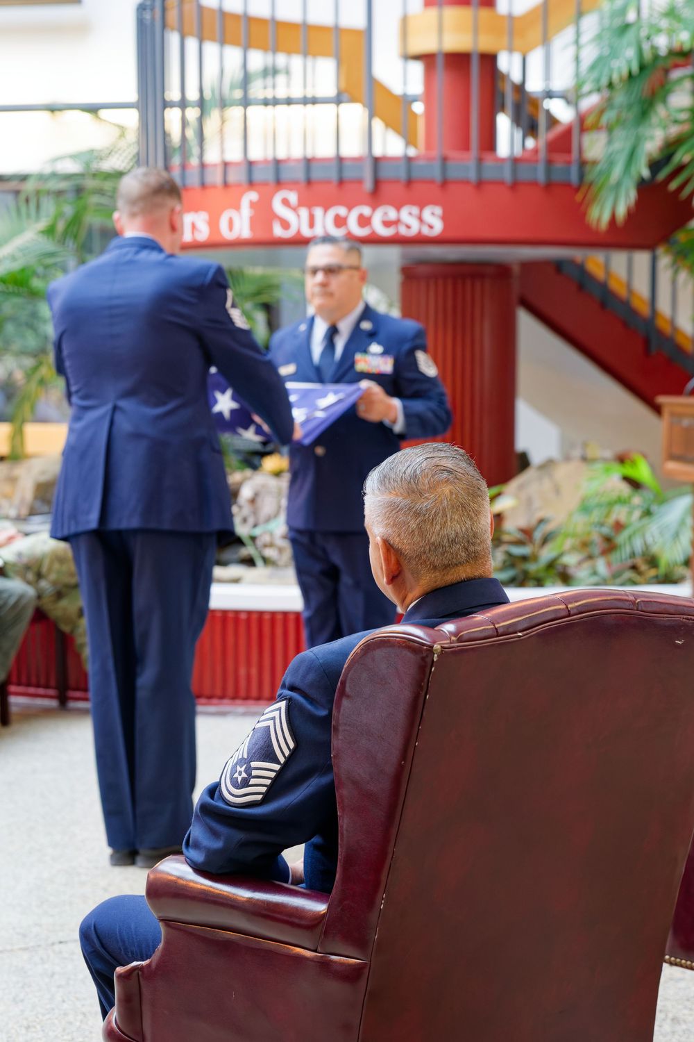 Air Force personnel perform a flag folding ceremony as a senior NCO watches from a leather chair.