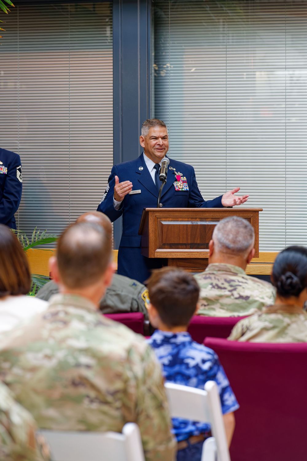 Military officer in dress blues speaks at podium before seated audience of uniformed personnel at official ceremony.
