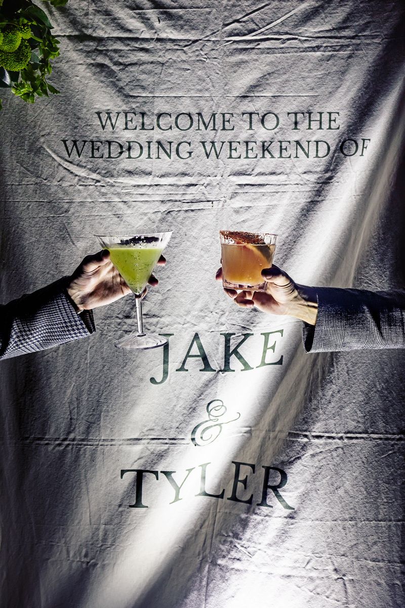 Two hands toasting cocktails in front of a wedding welcome banner for Jake and Tyler's wedding weekend.
