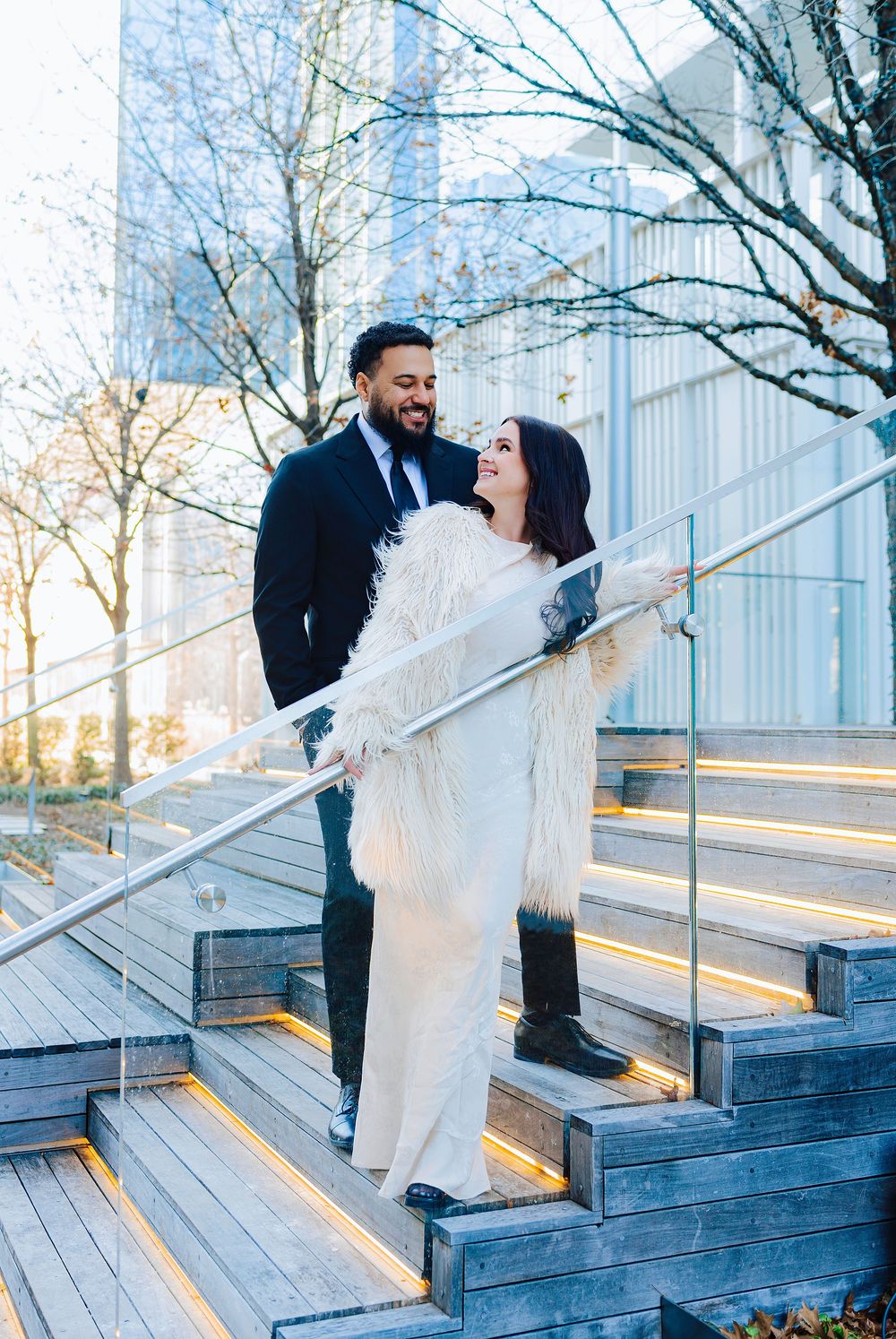 Couple in formal wear smiles on lit outdoor steps, woman in white gown with fur wrap, man in dark suit, urban setting.