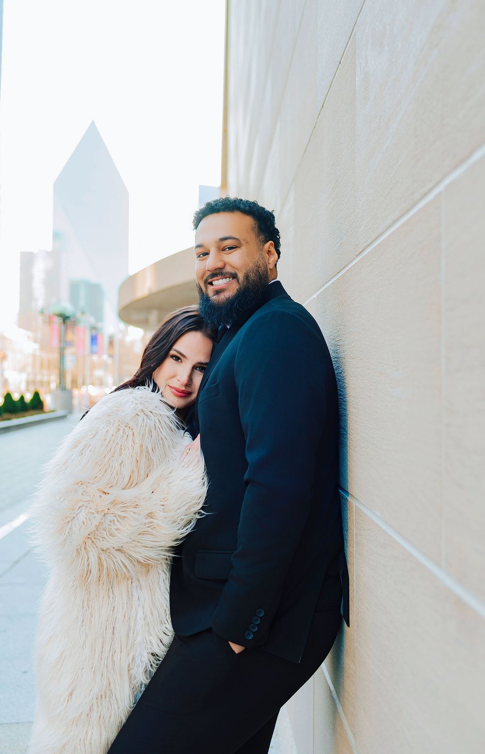 Couple posing against a wall in a city, woman in white fur coat leaning on man in black suit smiling.
