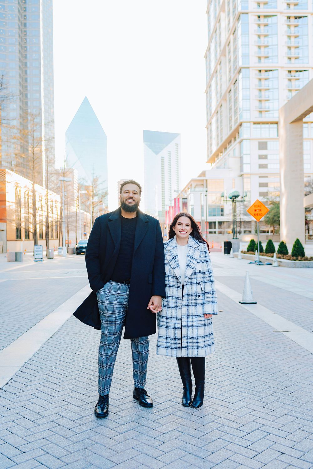 Couple holding hands in matching plaid outfits posing on urban plaza with Dallas skyline in background.