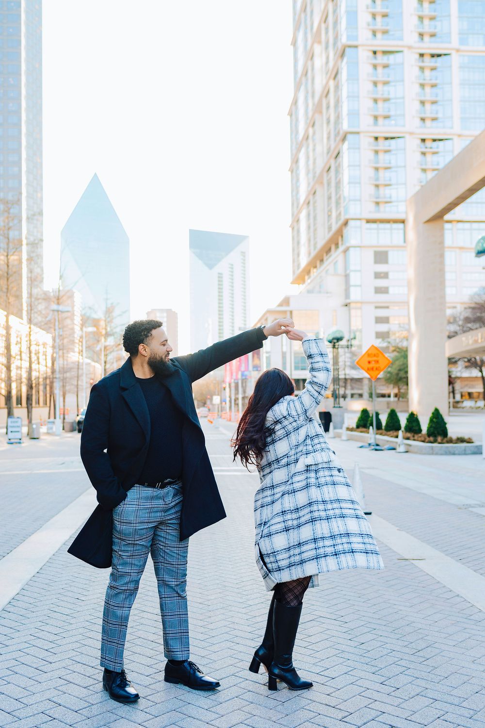 Couple dancing outdoors in urban setting, man in plaid pants and woman in checkered coat, city skyscrapers in background.