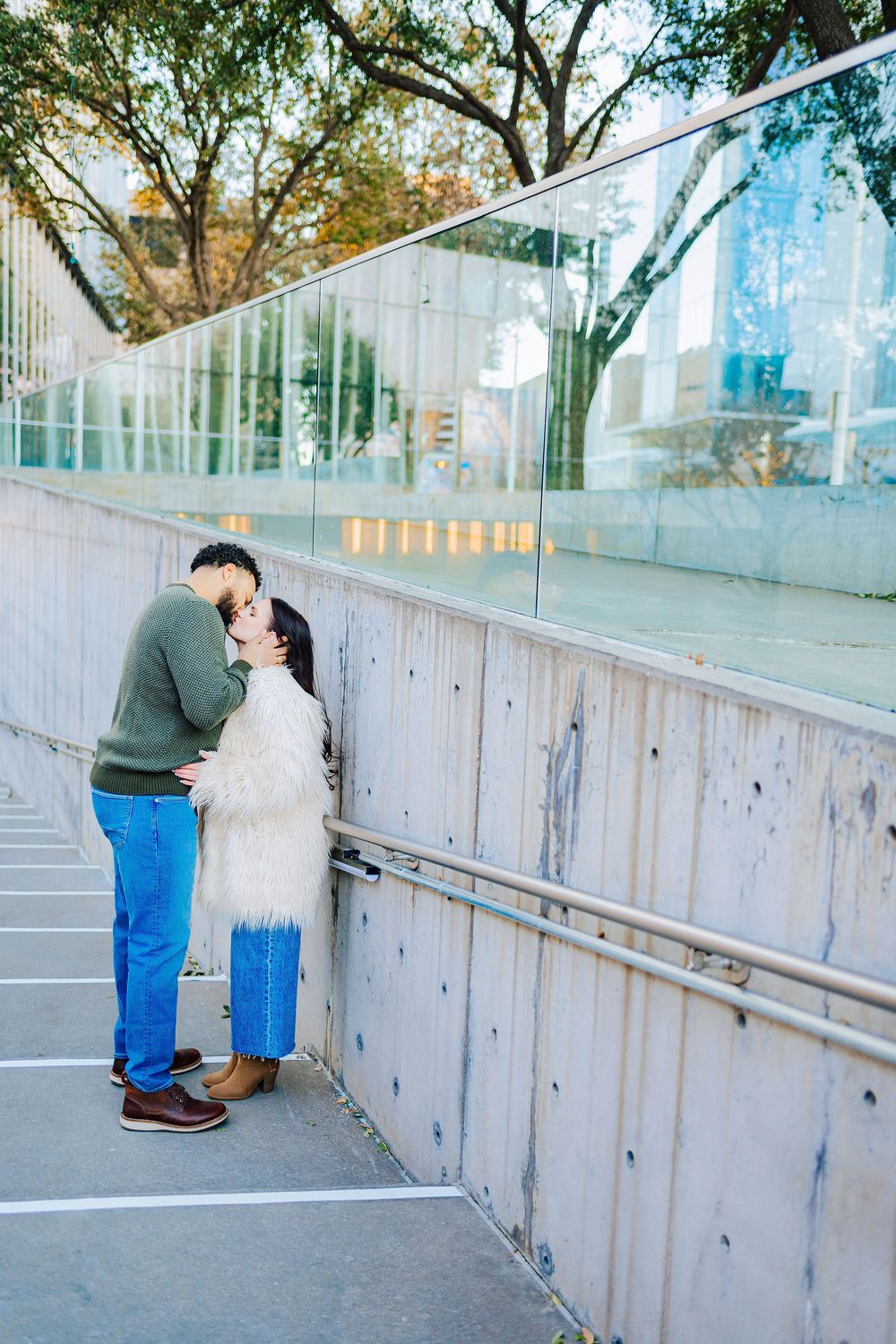 Couple sharing a romantic kiss on an urban walkway with glass walls and trees in the background.