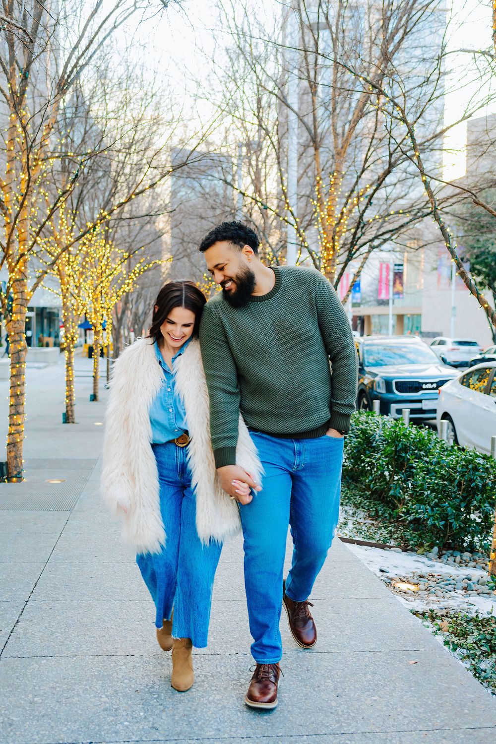 Couple walking hand-in-hand on a city sidewalk lined with fairy-lit trees in winter.