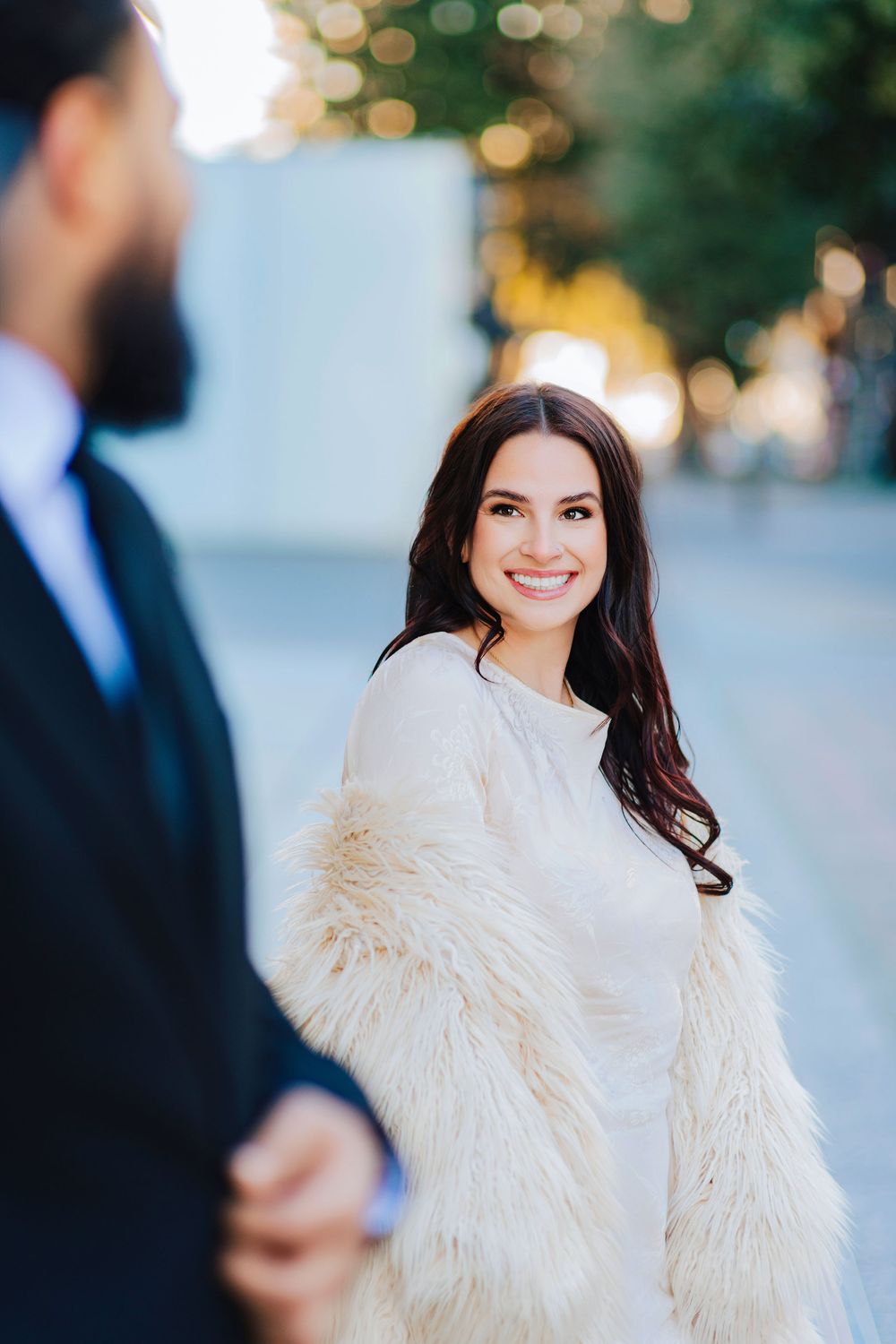 Smiling brunette woman in white fur coat holds hands with bearded man in black suit outdoors.