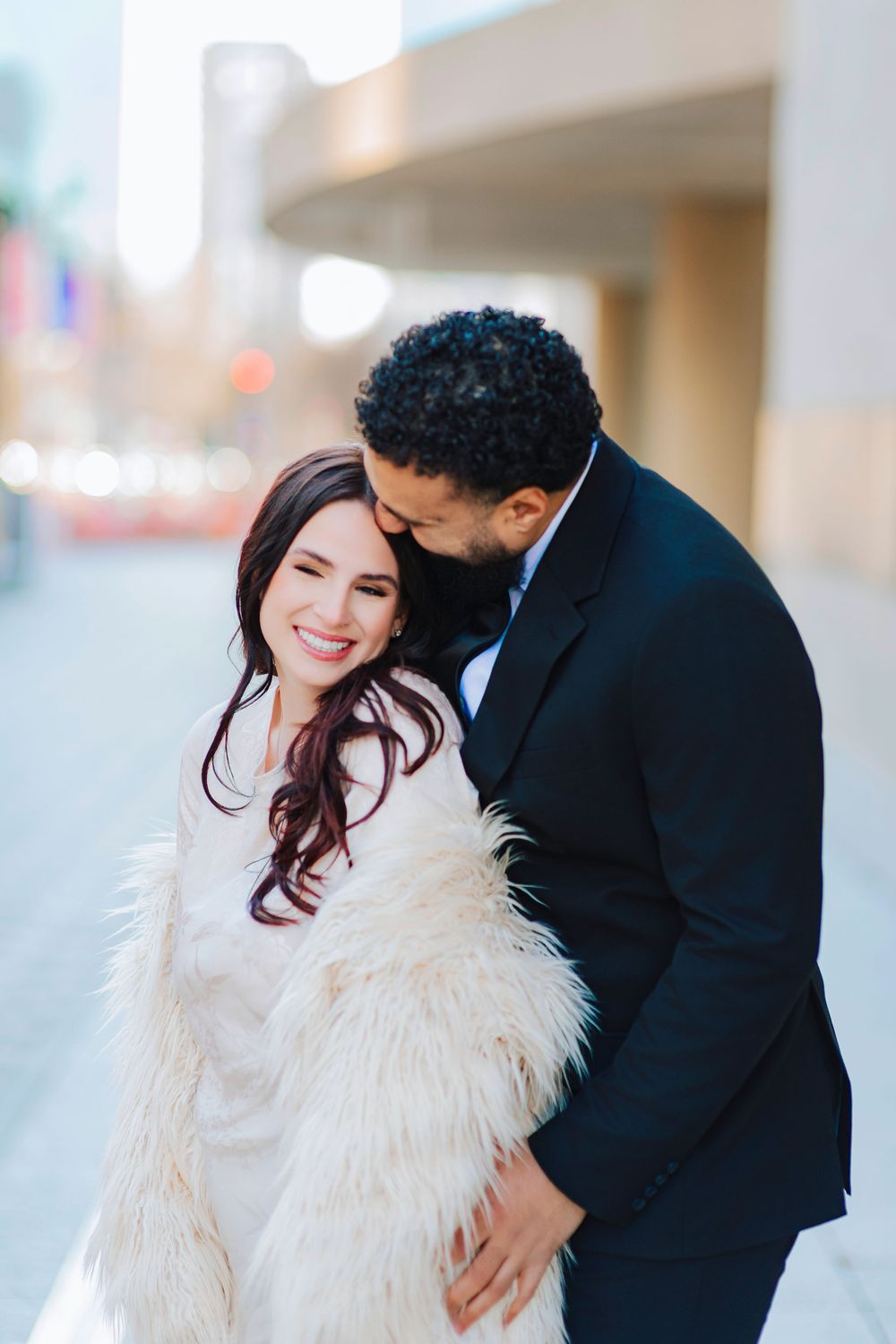 A smiling bride in a white fur wrap embraced by a groom in a dark suit on a city street.