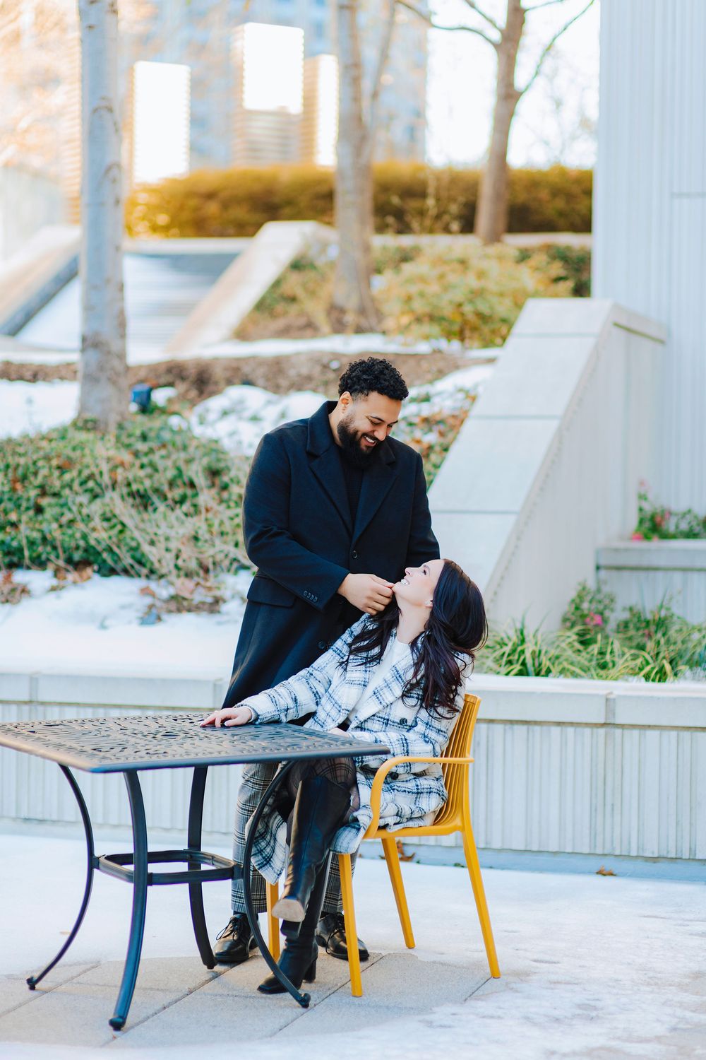 A couple shares a joyful moment at an outdoor table, man in black coat standing over seated woman in plaid jacket.