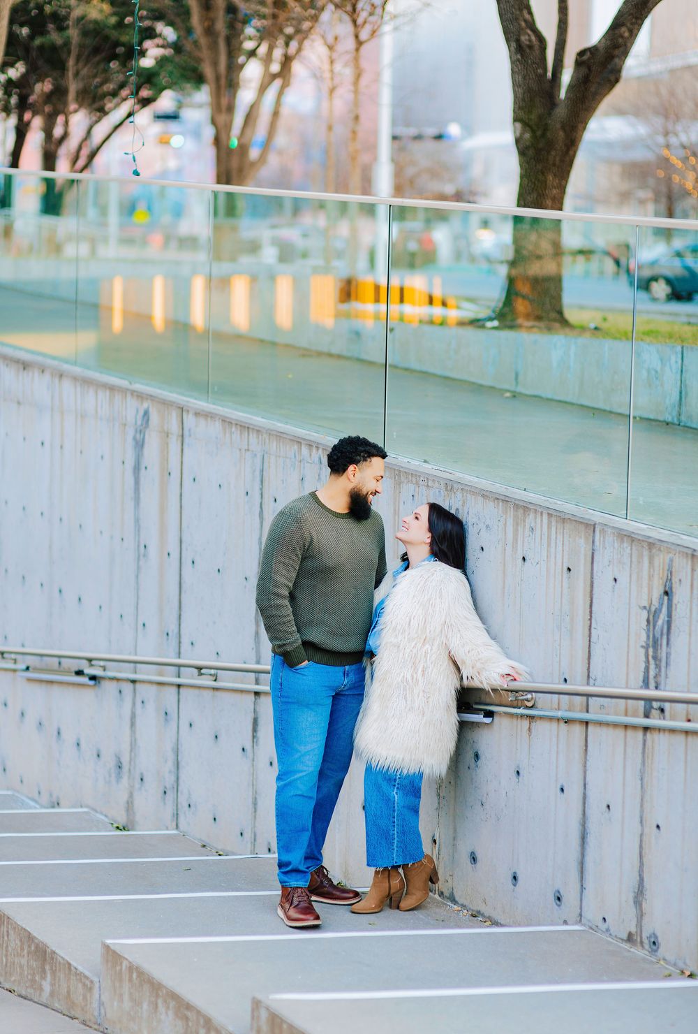 Couple sharing a romantic moment on concrete steps near a glass railing in an urban setting.