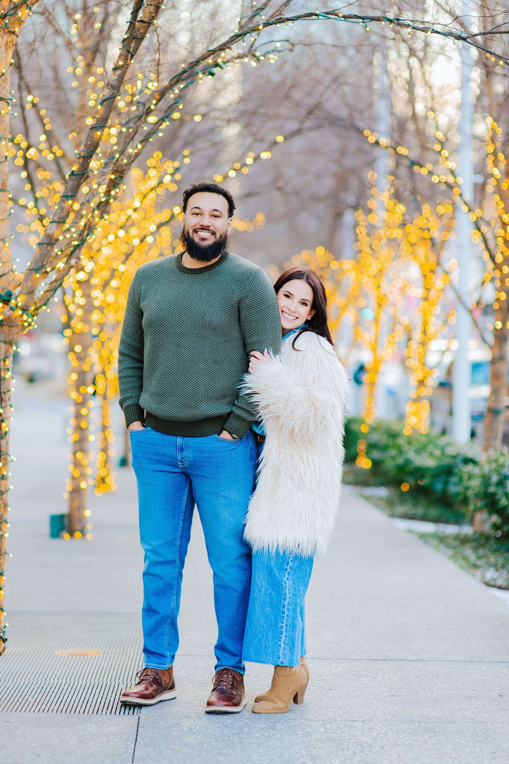 A smiling couple poses on a festive sidewalk lined with glowing yellow string lights on bare winter trees.
