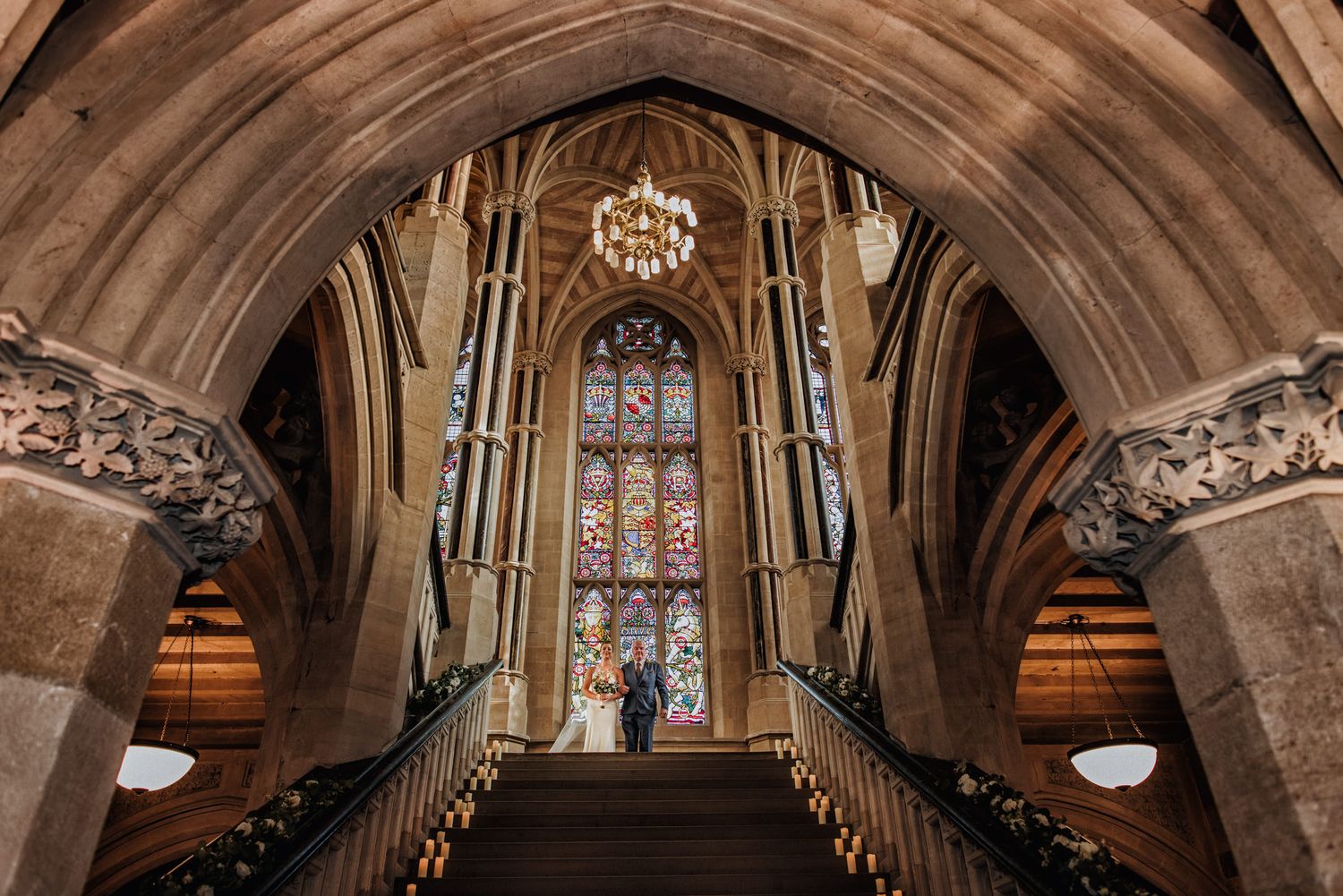 Couple stands at the top of a grand staircase beneath Gothic stone arches, with stained glass window and chandelier at Rochdale Town Hall