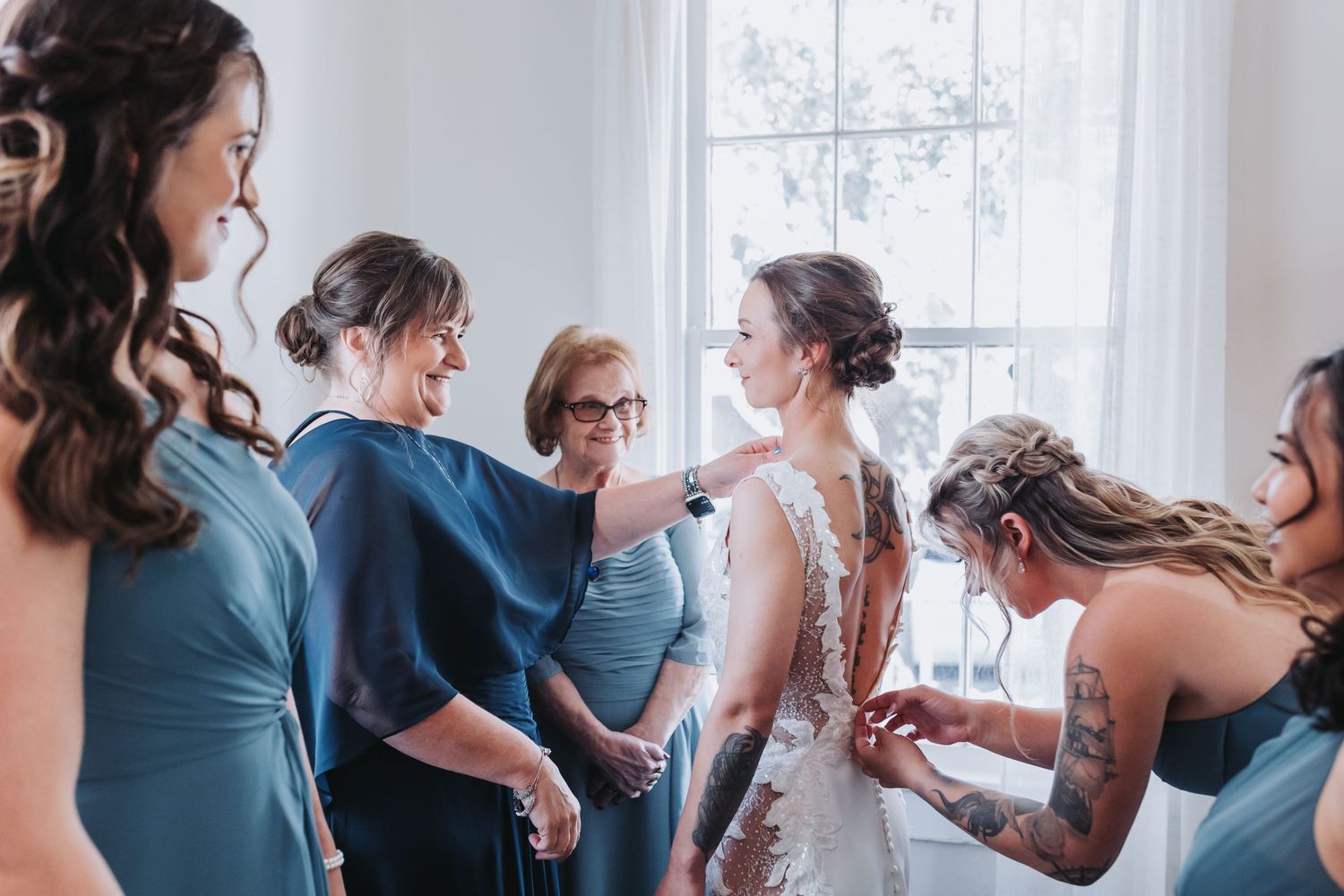 Bride in a lace wedding gown surrounded by bridesmaids in dusty blue dresses as they help her get ready by a bright window.