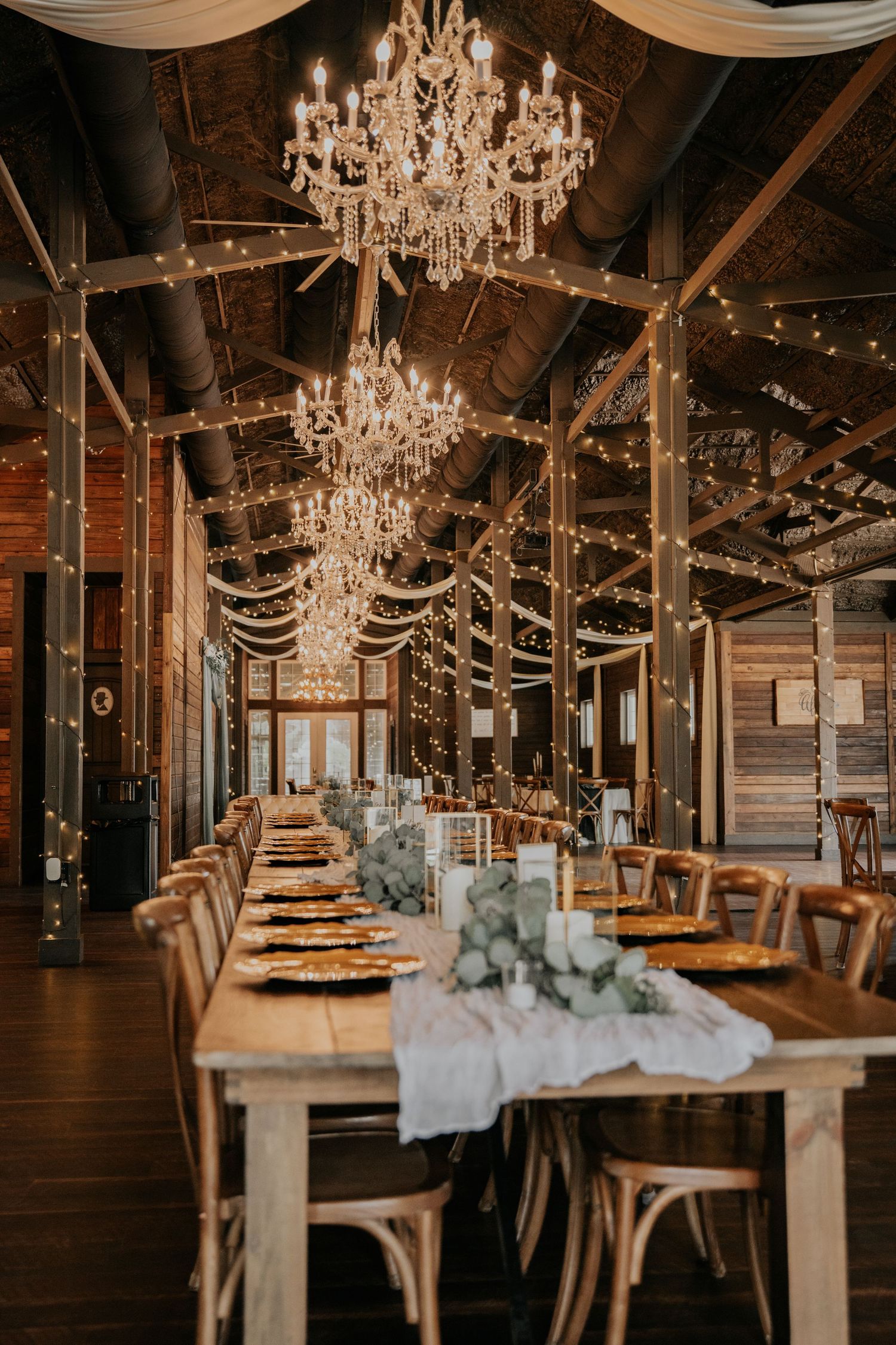 Rustic barn wedding venue with crystal chandeliers, string lights, and a long wooden farm table set with gold chargers and greenery.