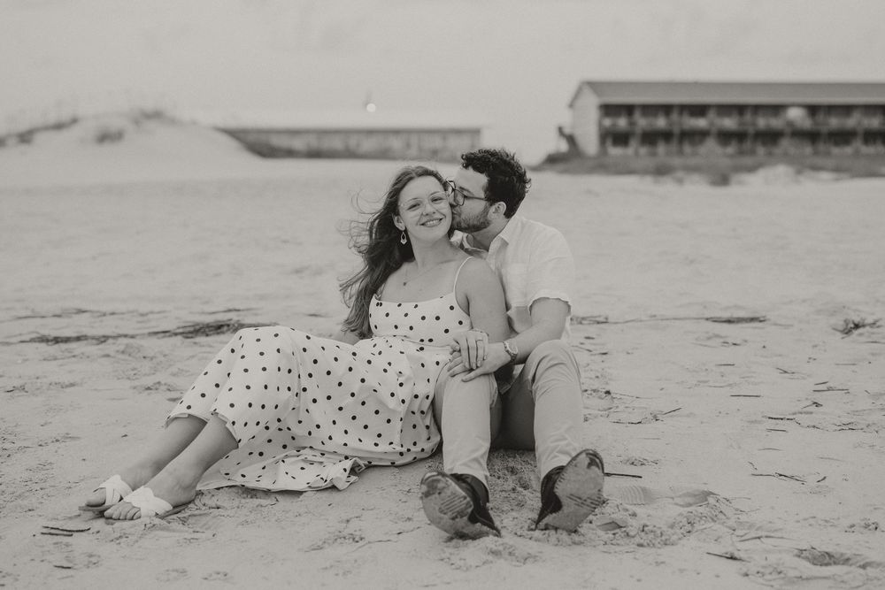 Black and white beach photo of a couple sitting on sand, man kissing woman's cheek, wearing polka dot dress.