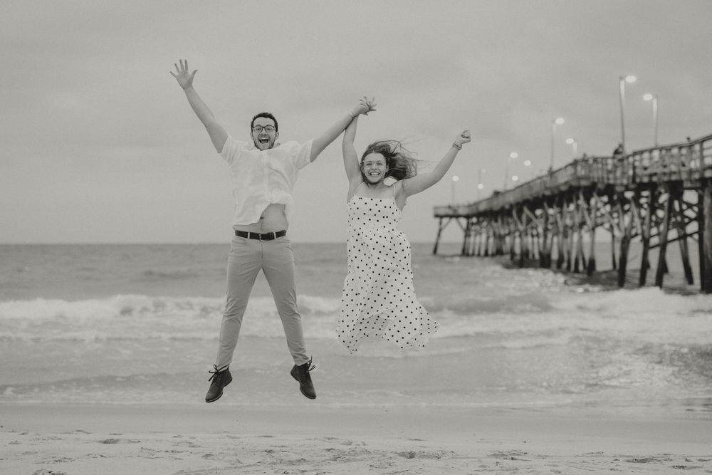 Joyful couple jumping on beach near pier in black and white photo.