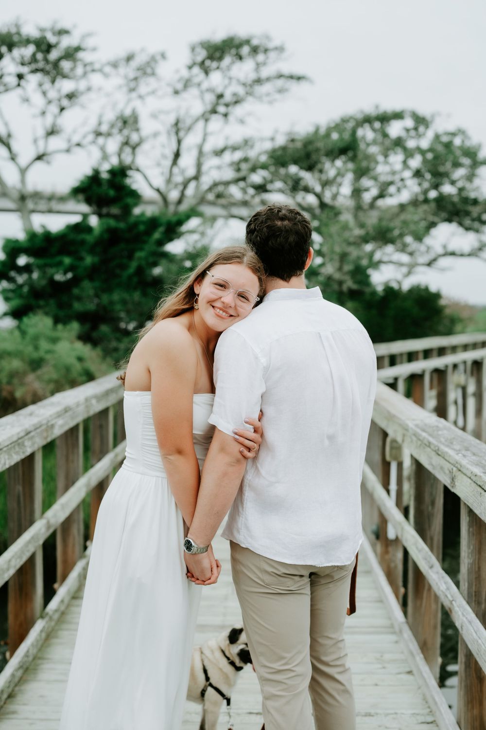 A smiling bride in a white dress embraces a man in a white shirt on a wooden bridge surrounded by trees.