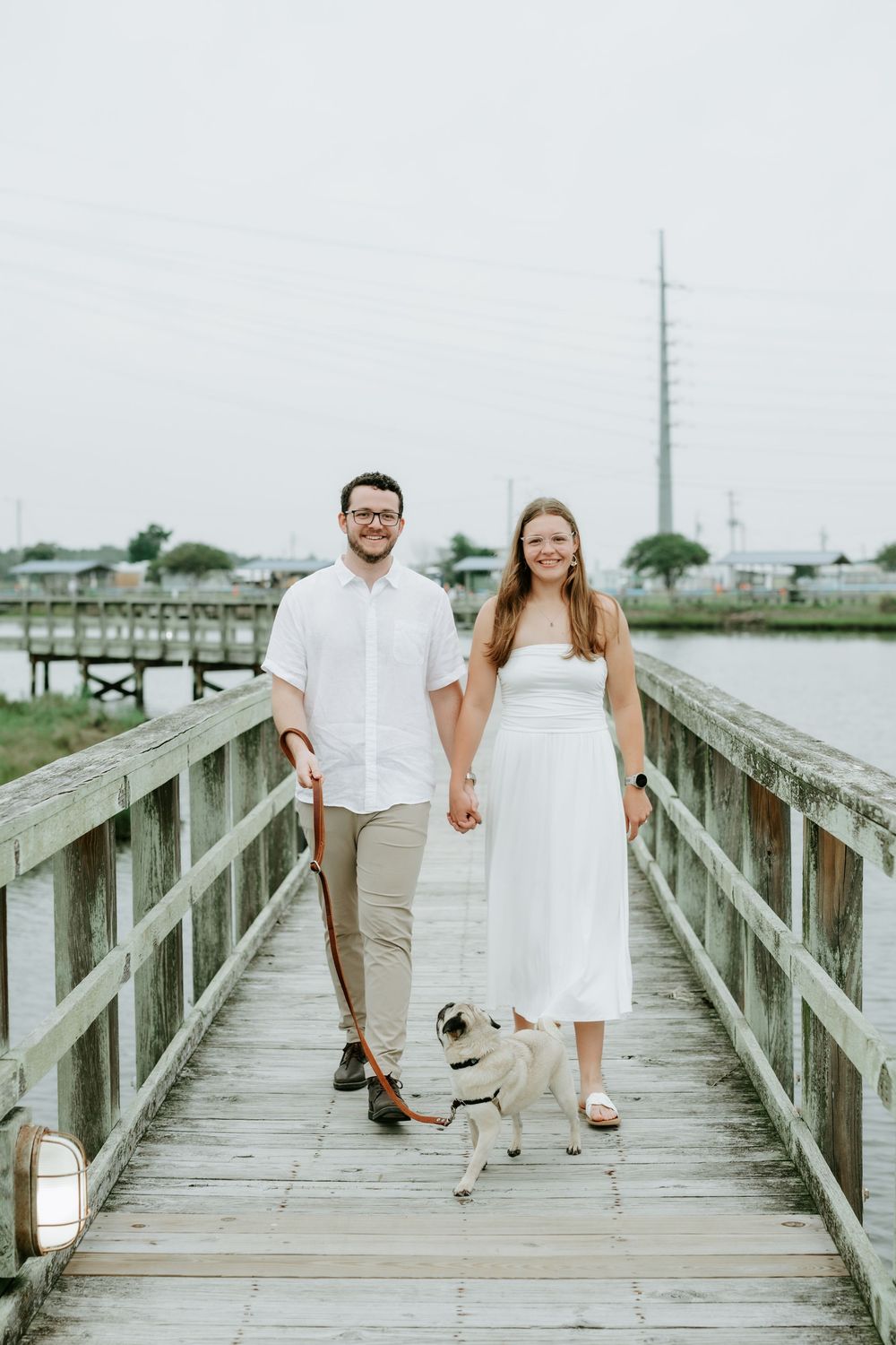 Couple in white outfits walking a pug on a wooden pier near the water on a cloudy day.