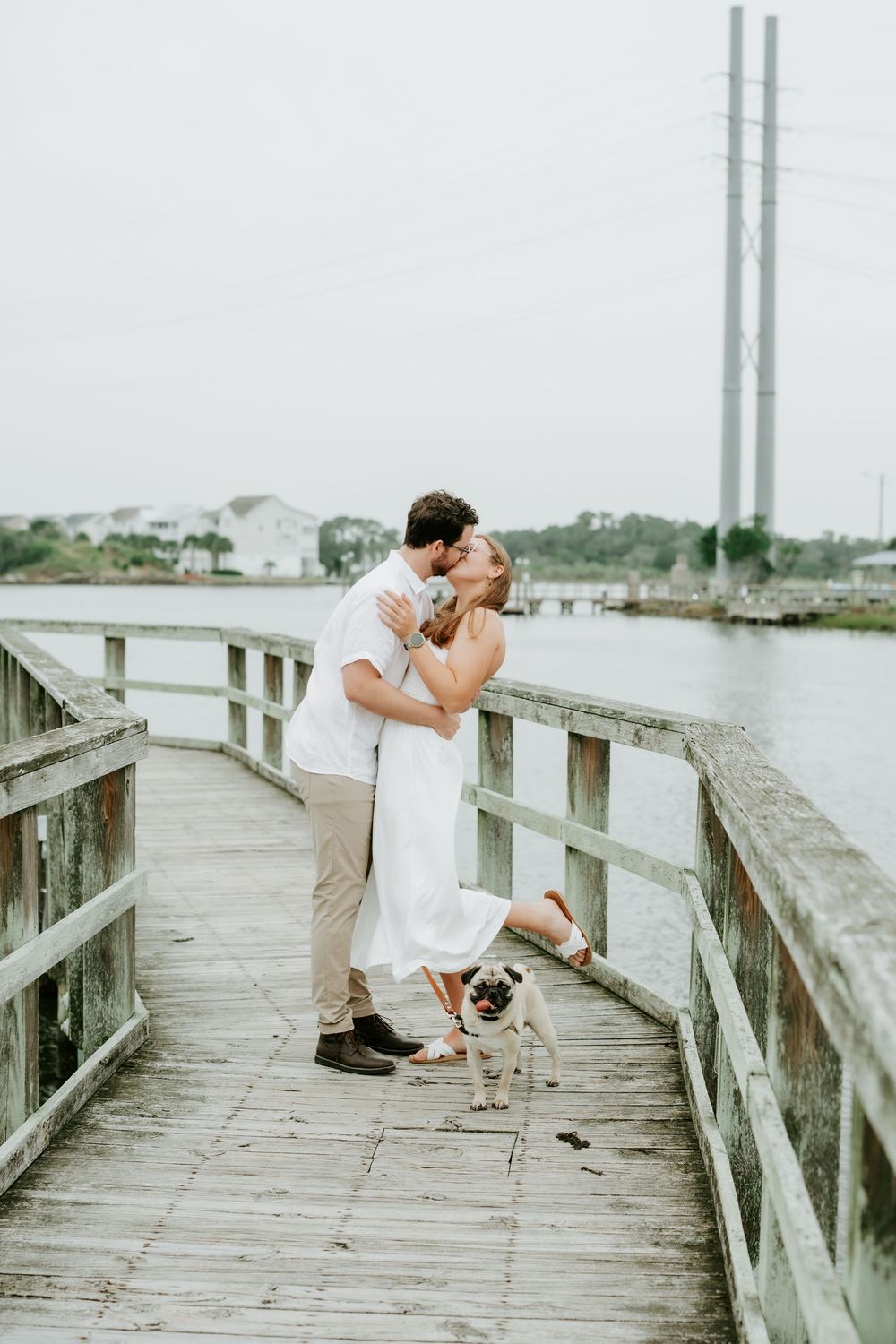 Couple kissing on a wooden pier with a pug dog, waterfront backdrop, romantic outdoor engagement photo.