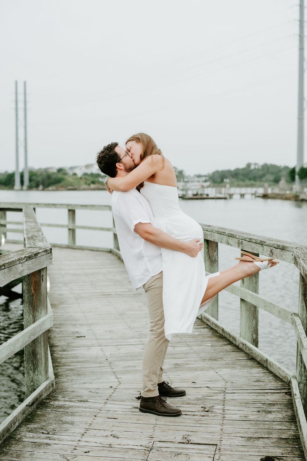 Couple sharing a romantic kiss on a wooden pier, man lifting woman in white dress, waterfront backdrop.