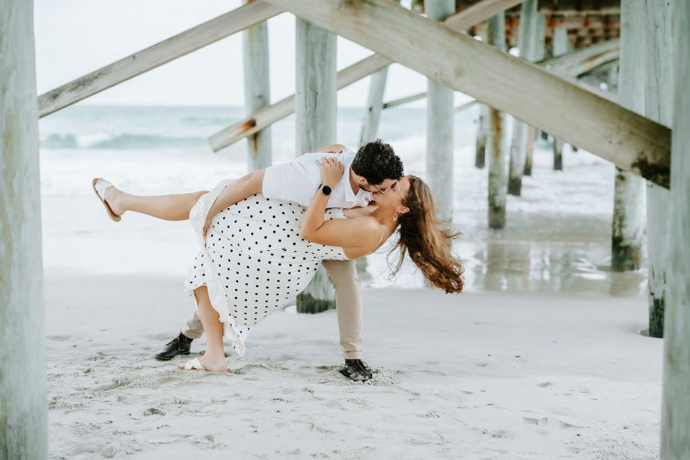 Couple sharing a romantic dip kiss on the beach near a pier, man in suit, woman in polka dot dress.