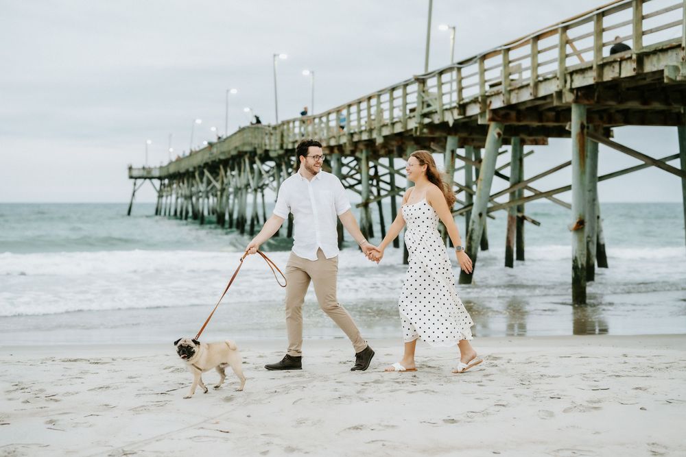 Couple walking a pug on a beach near a pier, holding hands, wearing casual summer outfits on a cloudy day.