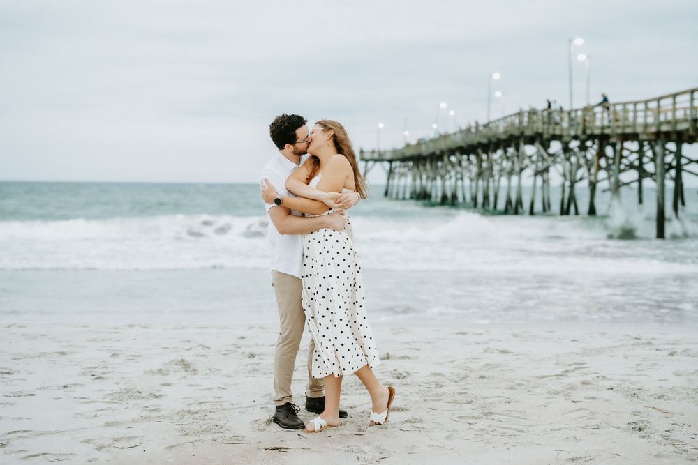 A couple shares a romantic kiss on a sandy beach near a pier, with ocean waves in the background.