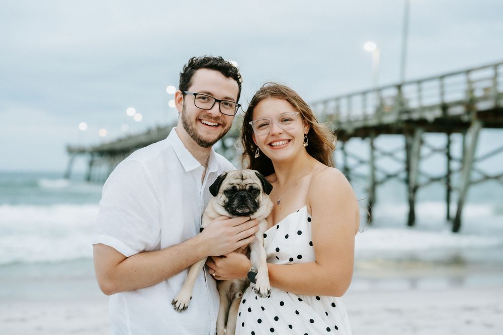 Couple smiling at beach pier holding a pug dog, woman in polka dot dress, man in white shirt, cloudy sky.
