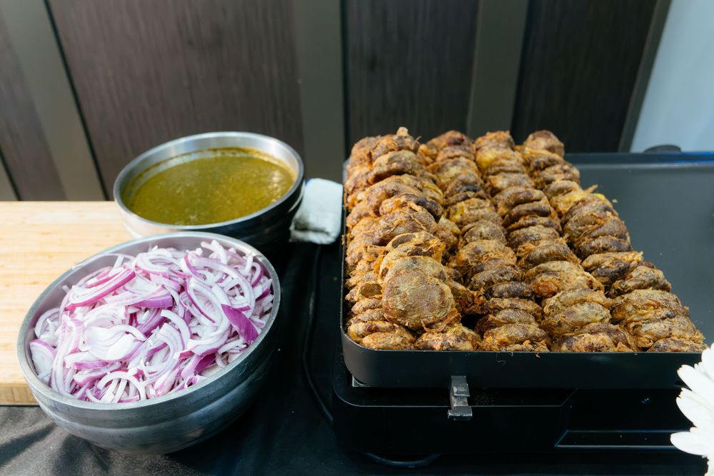 Tray of sliced roasted carnitas next to a bowl of sliced red onions and a pan of green salsa on a buffet table.