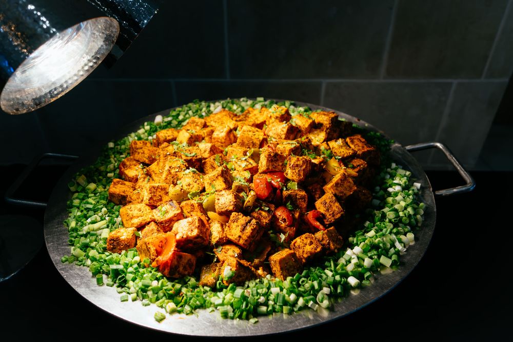 Spicy braised tofu cubes in red chili sauce topped with chopped green onions, served on a large round metal tray under warm buffet lighting.