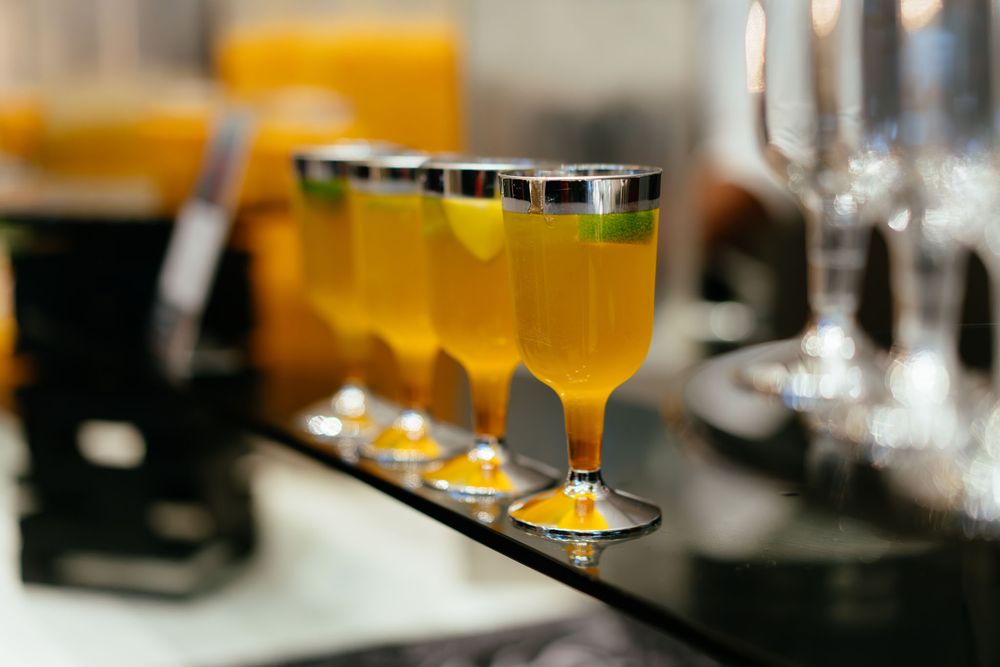 Row of stemmed plastic cups filled with orange juice and topped with lime slices on a black bar counter with blurred glasses behind.