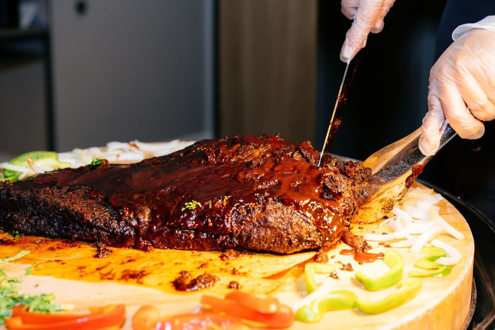 Gloved hands carve a glazed barbecue brisket on a wooden board with sliced onions, peppers, and fresh garnishes.