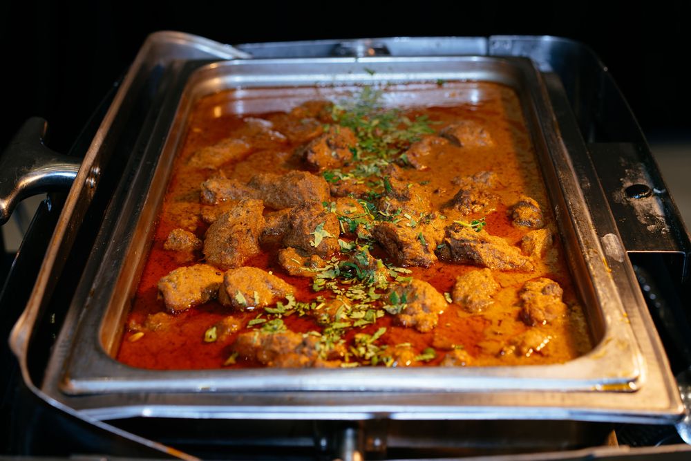 Chunks of meat simmered in a rich red curry sauce and garnished with fresh herbs in a stainless steel chafing dish.